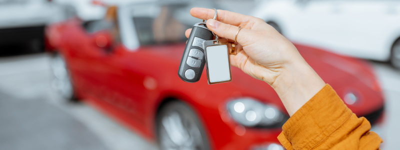 A hand holding car keys with a red car blurred out in the background, symbolising new car ownership.