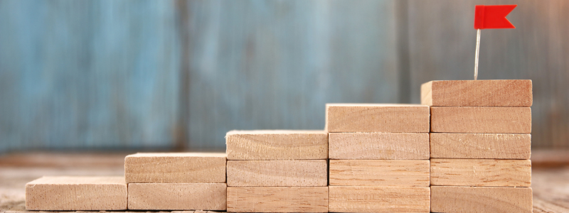 Wooden blocks rising in number from left to right with a flag on the end block, representing a step by step journey.