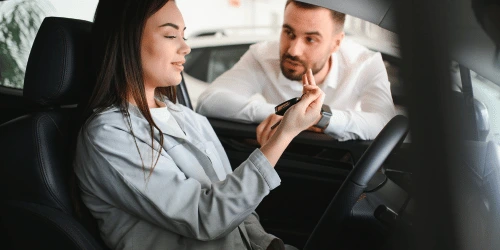 A woman talking to a man whilst sat in a car, likely in a car showroom.